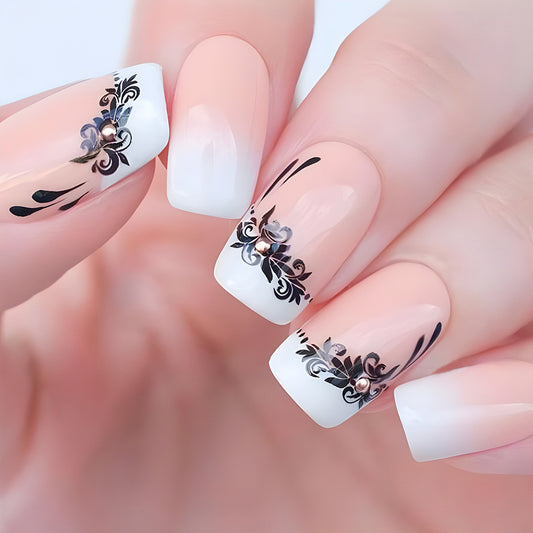 A set of press-on nails showcasing a medium length coffin shape with a pink to white ombre design and black floral accents.