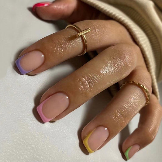 A person's fingers showcasing press-on nails with a beige, short, square French tip design, with pastel-colored accents on the corners of the nails.