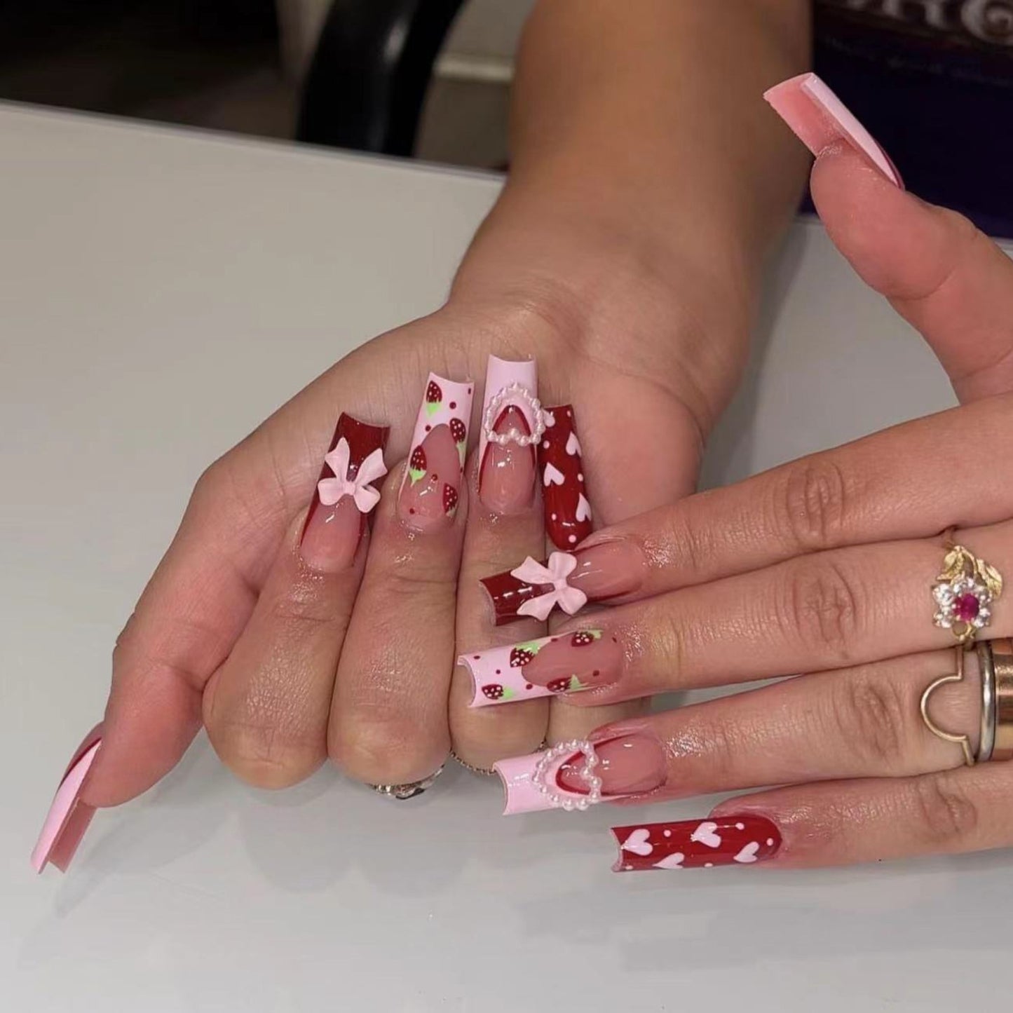 A set of press-on nails featuring long coffin-shaped nails in red with white dots and floral patterns, adorned with small pink flowers and gold accents.