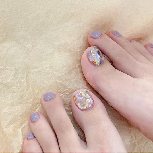 A close-up image of a person's toes with round, short fingernails featuring iridescent gem accents, painted in a lavender color.