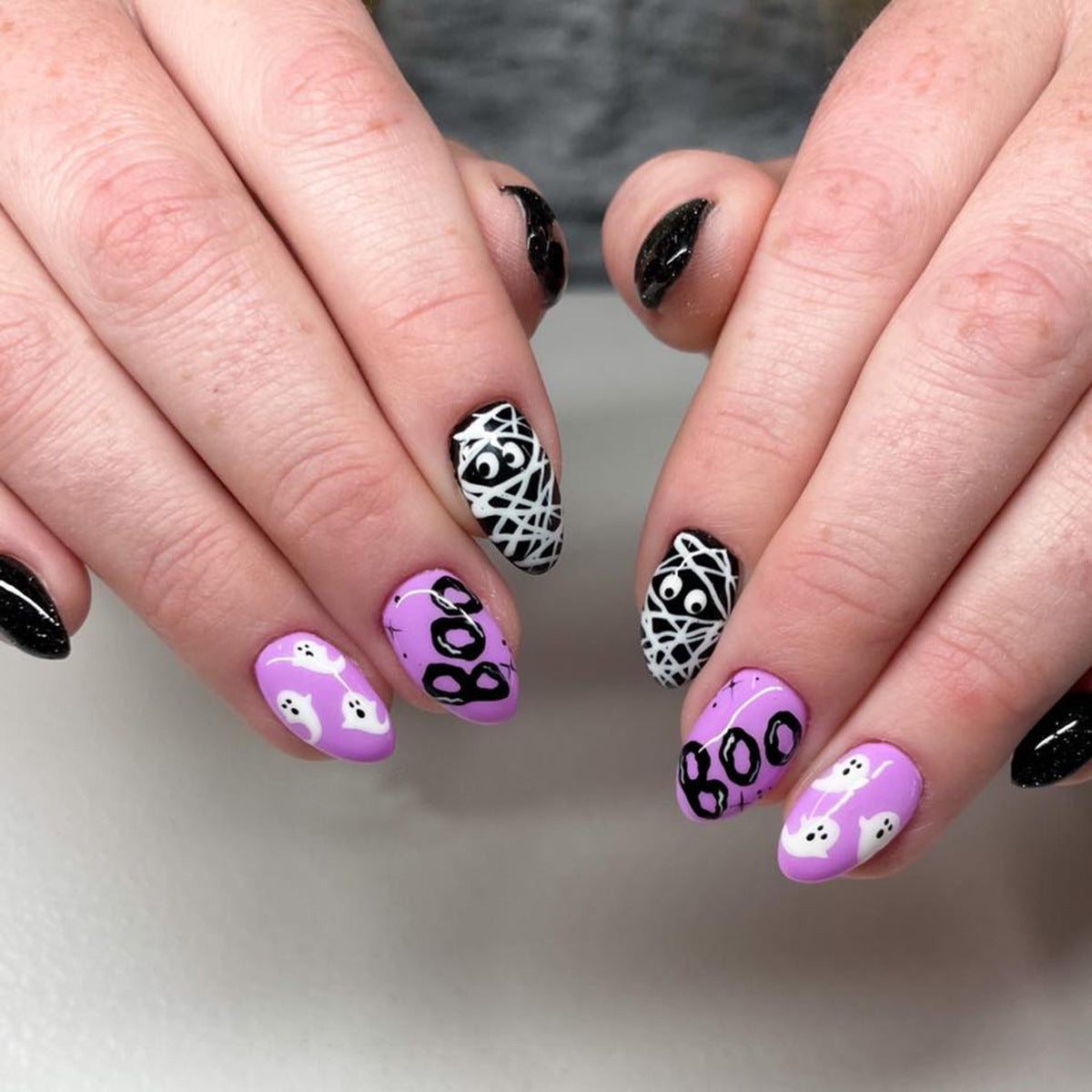 A person's hands displaying press-on nails with a Halloween theme, featuring purple nails with 'BOO' text and black and white patterned nails.
