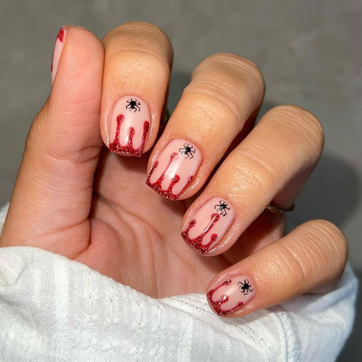 A hand with press-on nails featuring a red glitter design with black spider accents and a simulated blood drip effect, indicative of a Halloween theme.