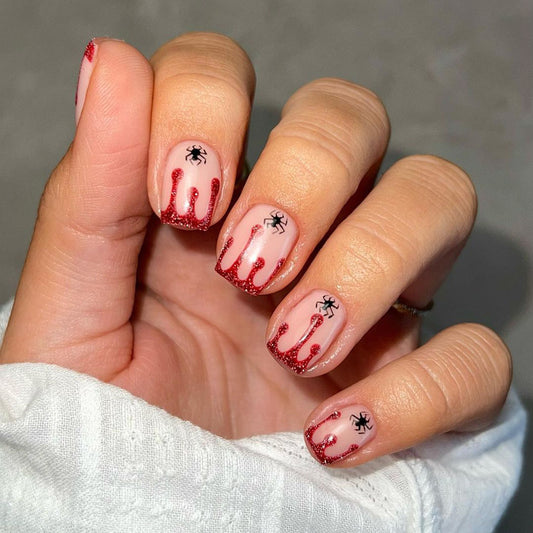 A hand with press-on nails featuring a red glitter design with black spider accents and a simulated blood drip effect, indicative of a Halloween theme.