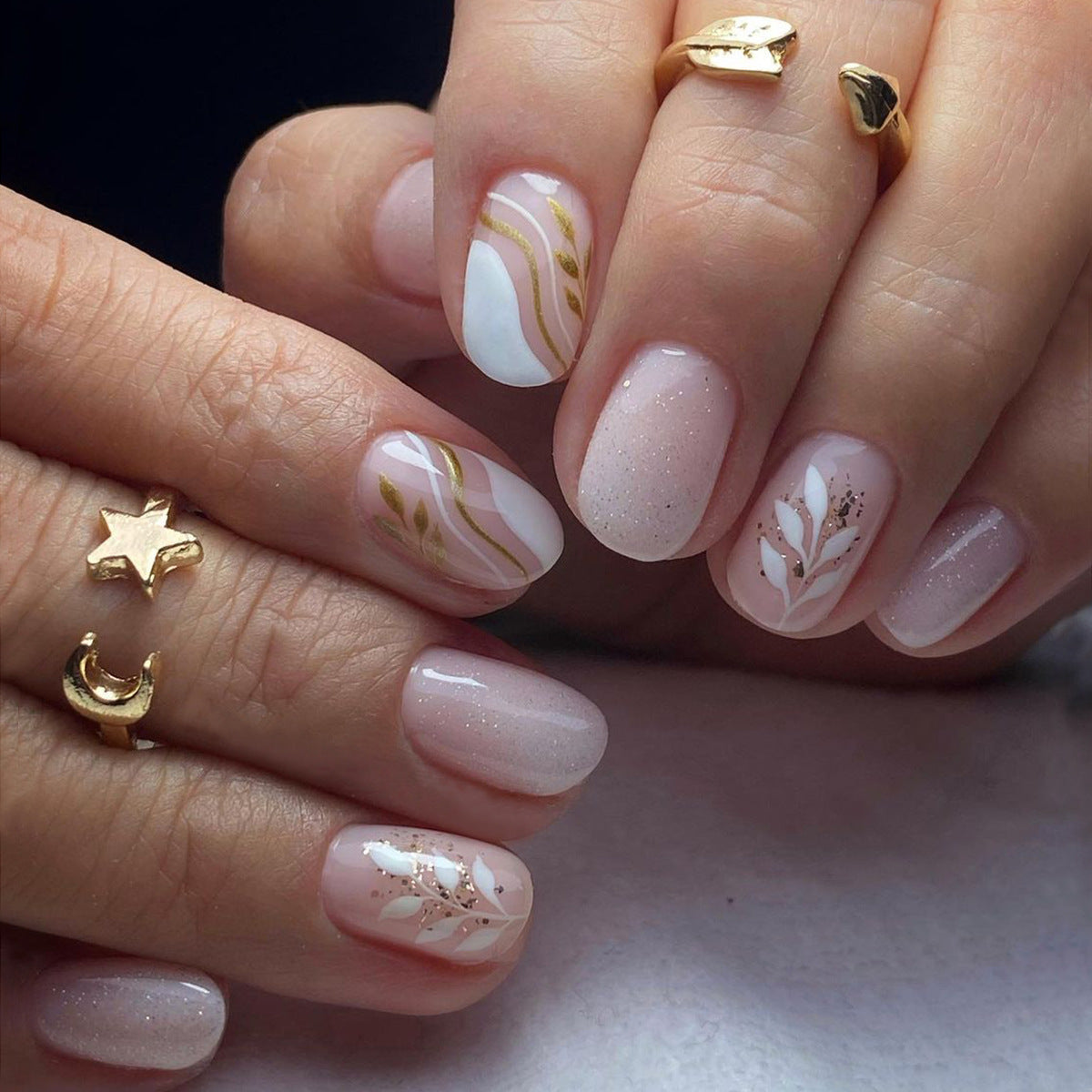 A set of short, round gold press-on nails with a floral pattern on a person's fingers, accompanied by gold rings.