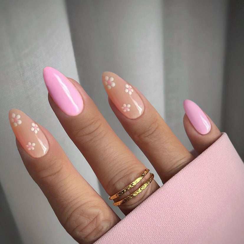 A hand with pink almond-shaped press-on nails with floral accents, wearing a pink ring, held in a pink case.