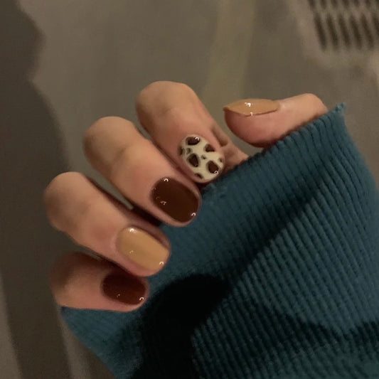 A hand with short square nails painted in brown shades, featuring a patterned nail art.
