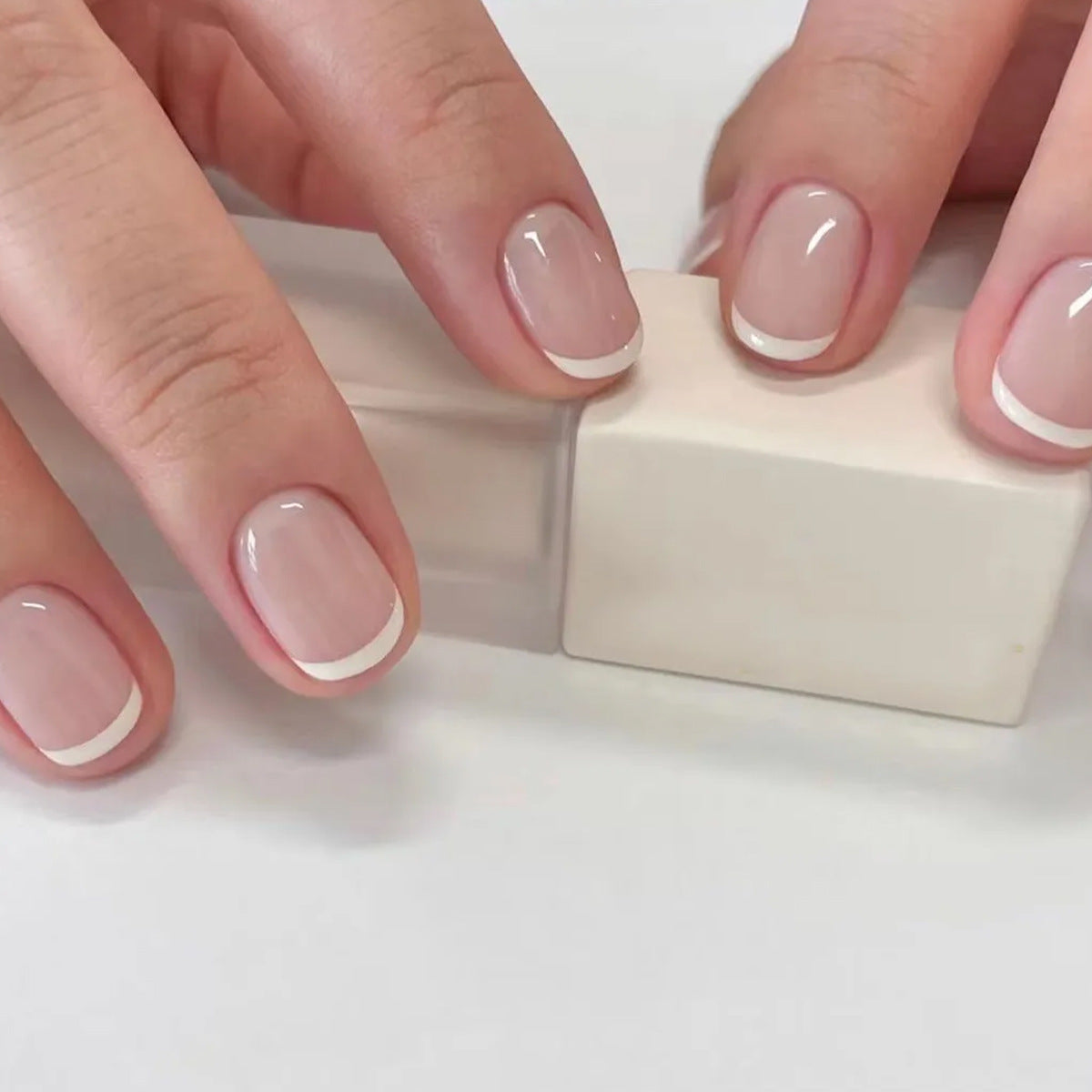 A close-up image of fingernails with white French tips using press-on nails, resting on a white surface.