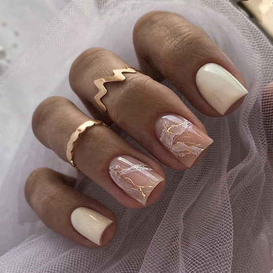 A set of press-on nails featuring a beige marble design on short, square-shaped nails. Accompanied by gold rings on the fingers.