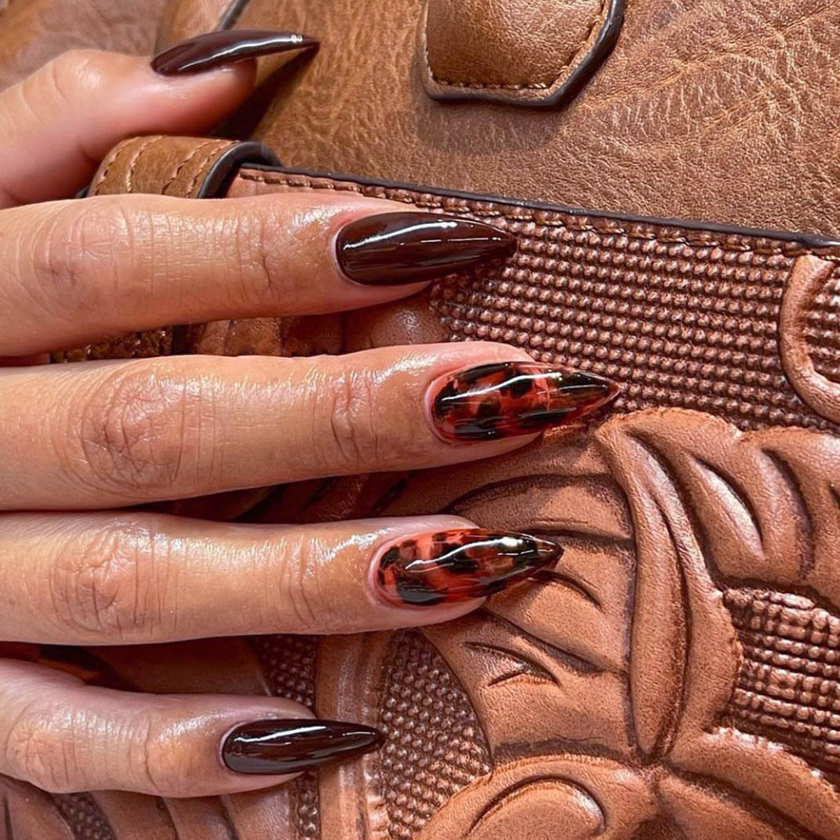Close-up of hands with tortoiseshell nail design holding a brown leather bag.