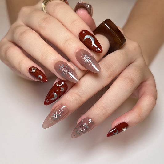 A person's hands showcasing press-on nails with a medium almond shape. The nails are painted in a red shade with a glossy finish, decorated with silver geometric patterns.