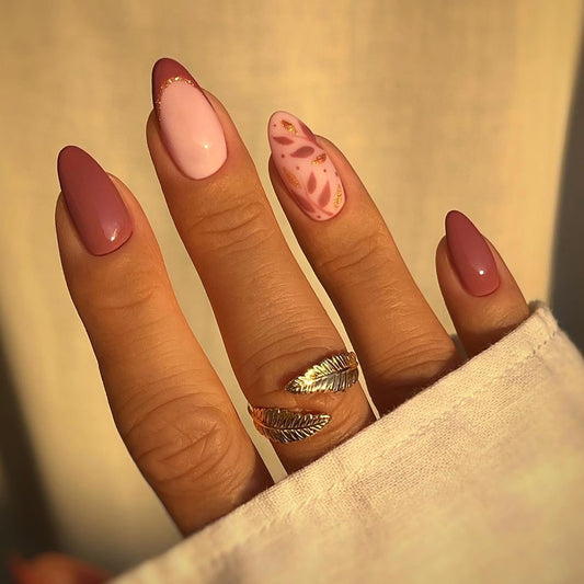A hand with medium almond brown false nails featuring a leaf pattern on the pinky nail.