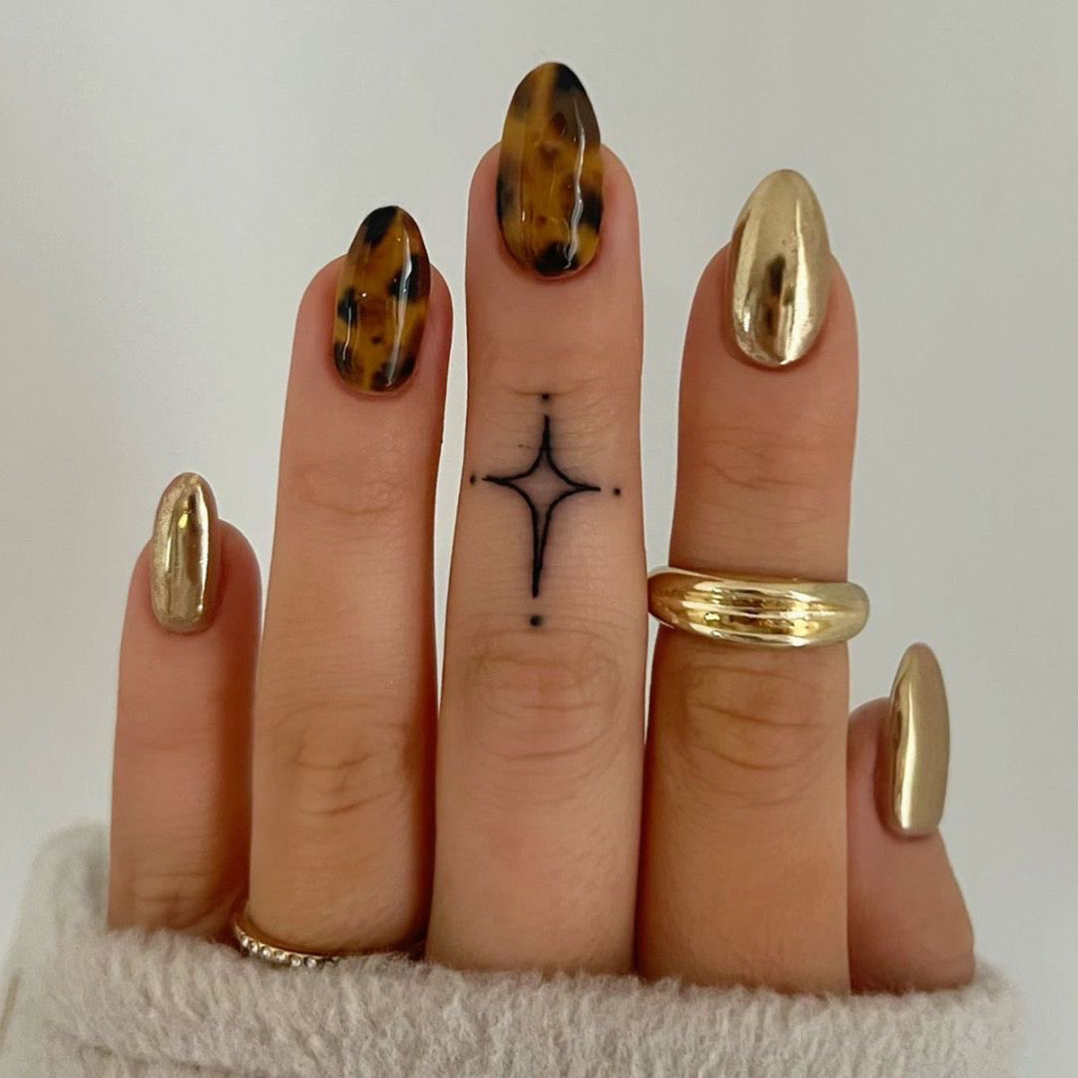 A set of press-on nails featuring a medium almond tortoiseshell pattern with metallic accents and a single ring on the finger.