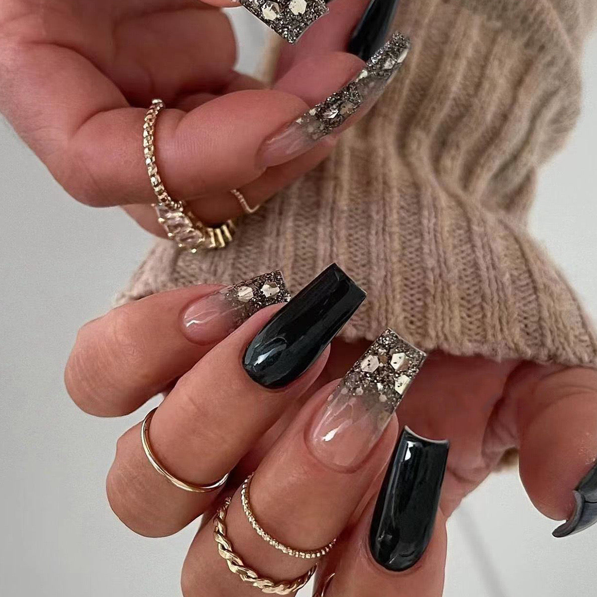 A close-up image of a person's hand with long black press-on nails featuring gold flake overlays, wearing gold rings, and a beige sweater.