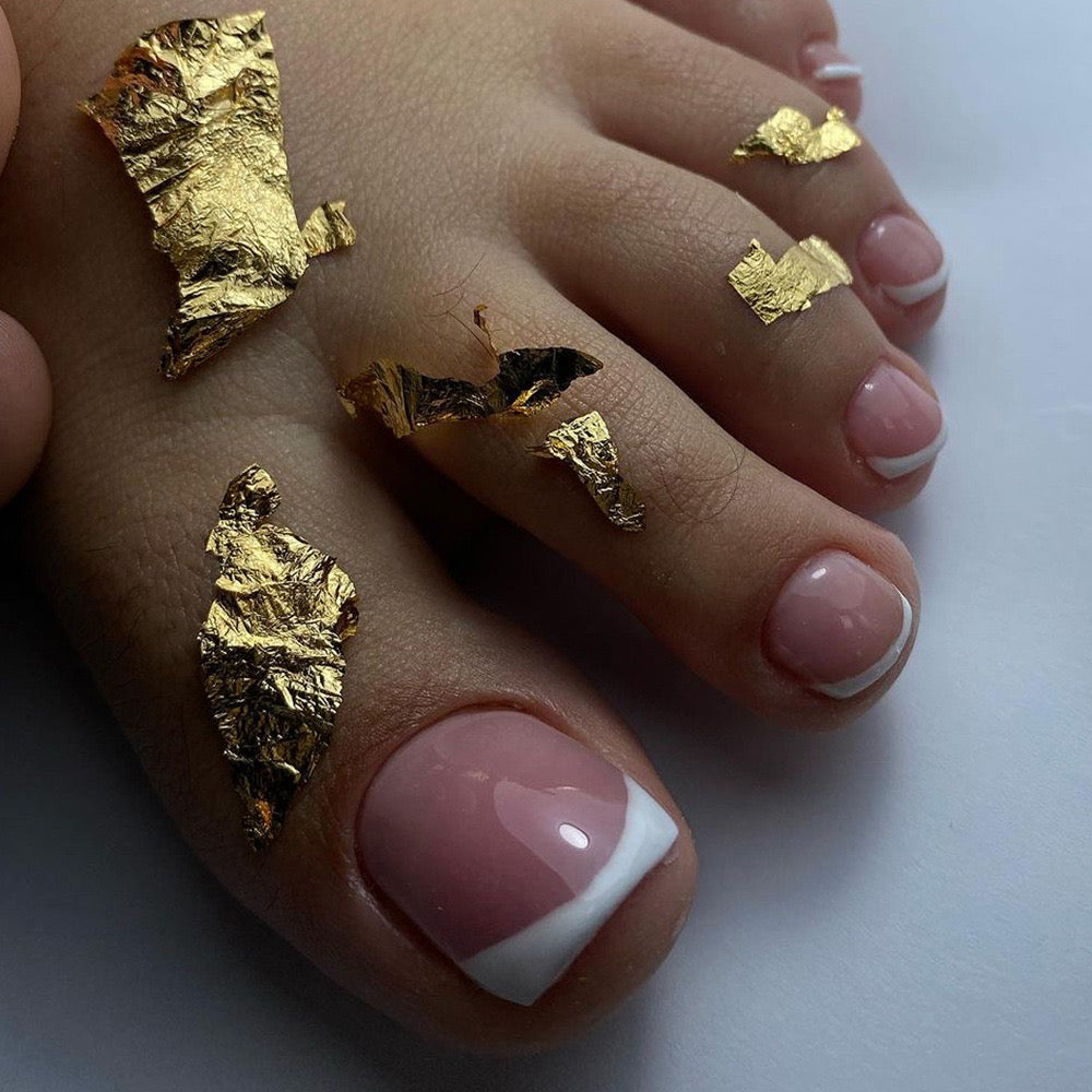 A close-up image of press-on toenails with a soft pink color, white tips, and gold leaf accents.