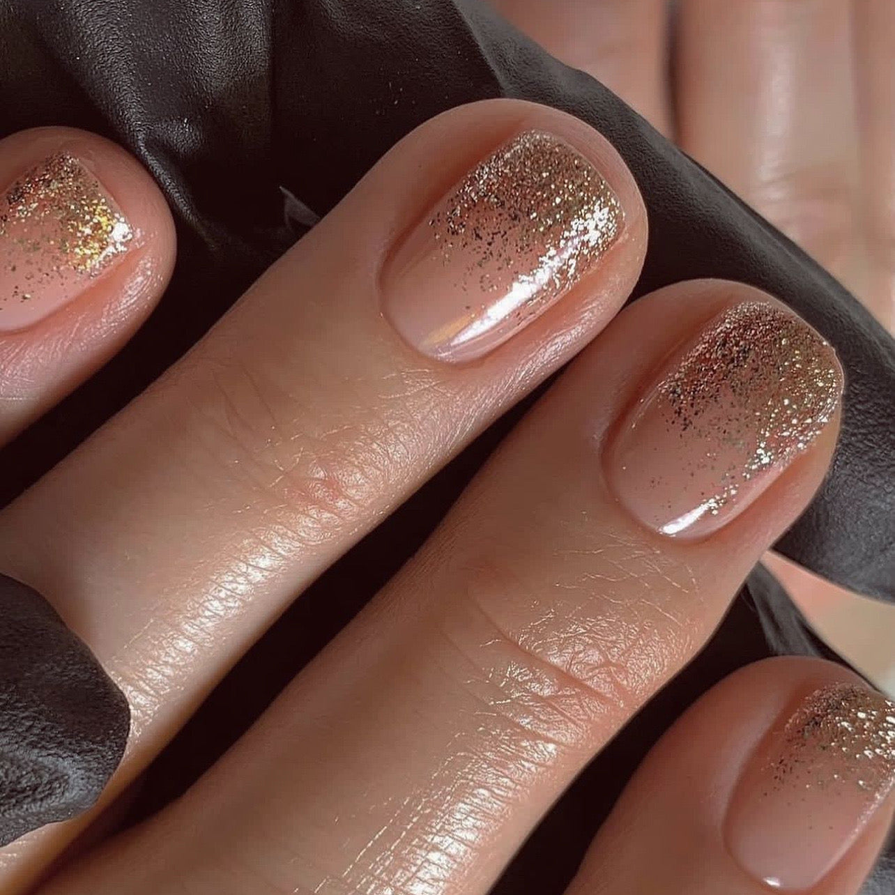A set of beige press-on nails with gold glitter gradient, displayed on a hand against a black background.