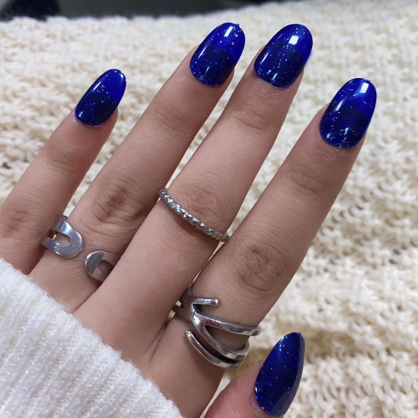 A person's hand with deep blue round-shaped press-on nails with glitter accents, wearing a silver ring and a light-colored sweater.