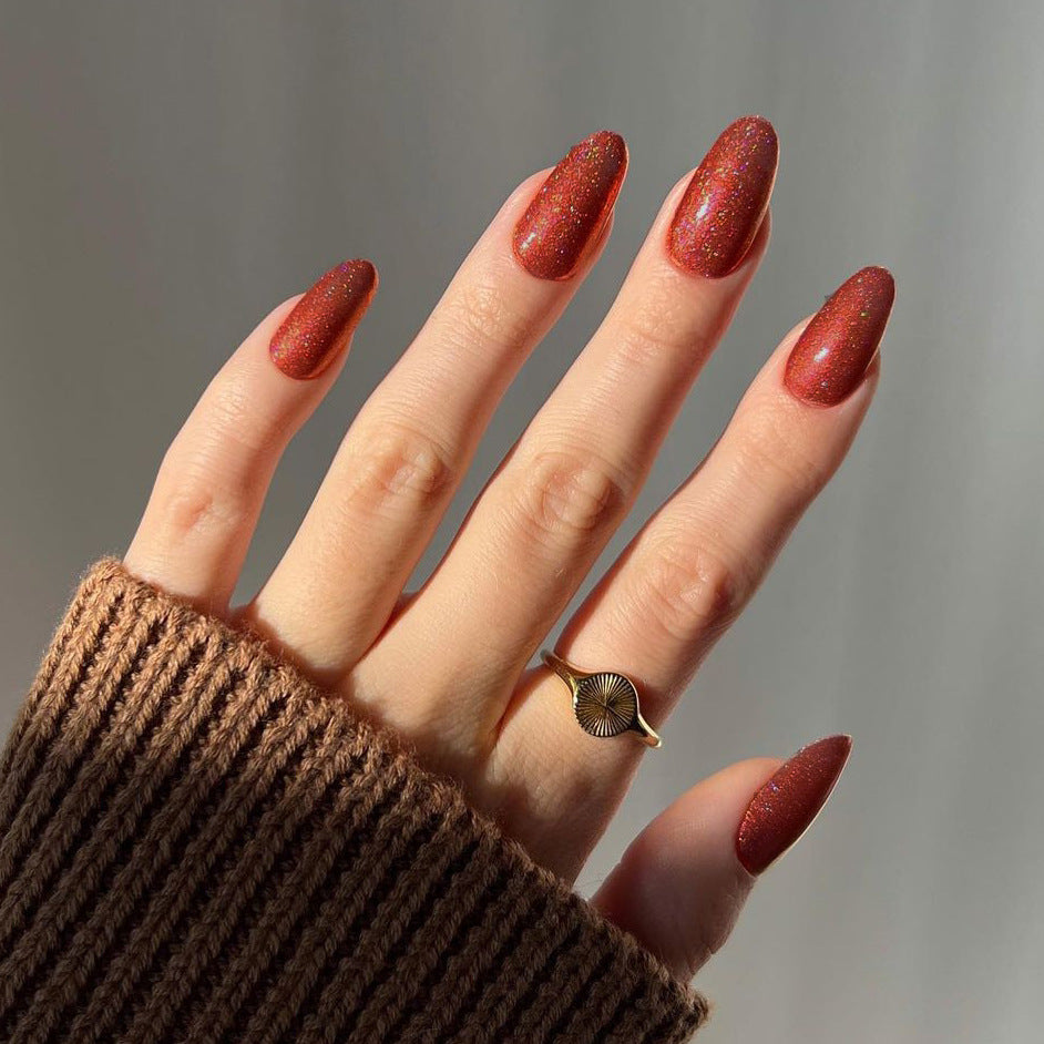 Hand with red glittery nails wearing a brown sleeve and gold ring on a neutral background