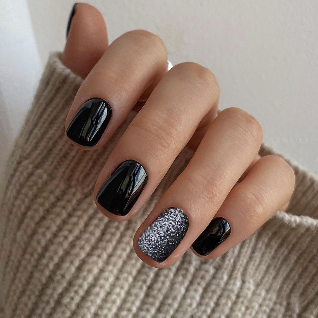 A hand with medium length squoval shaped black press on nails with one nail featuring a glitter accent.