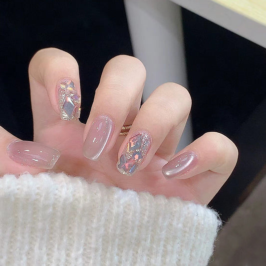 A hand with beige press-on nails featuring square shapes and multicolored glittering accents, alongside rose gold-colored flakes.
