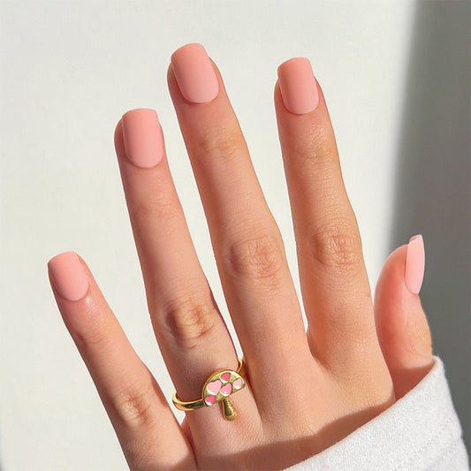 A hand with short square pink nails with green accents, wearing a gold ring with a pink gem.