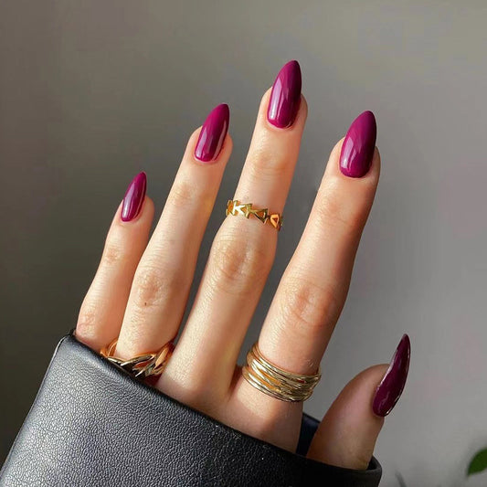 A hand with long almond-shaped pink glossy press-on nails, wearing multiple gold rings, holding a black object.