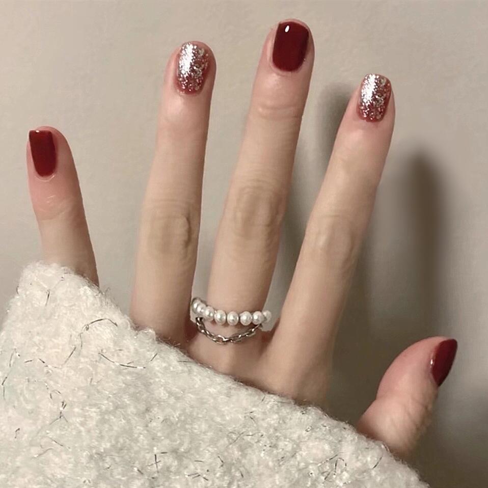 A hand with short squoval red nails with glitter, wearing a white pearl ring, resting on a white textured surface.