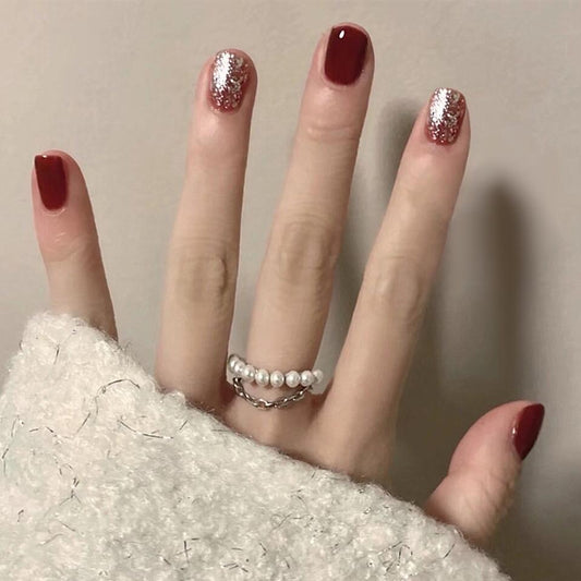 A hand with short squoval red nails with glitter, wearing a white pearl ring, resting on a white textured surface.