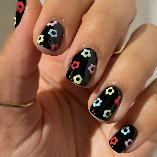A hand showcasing press-on nails with a black base and decorated with small multicolored floral designs.