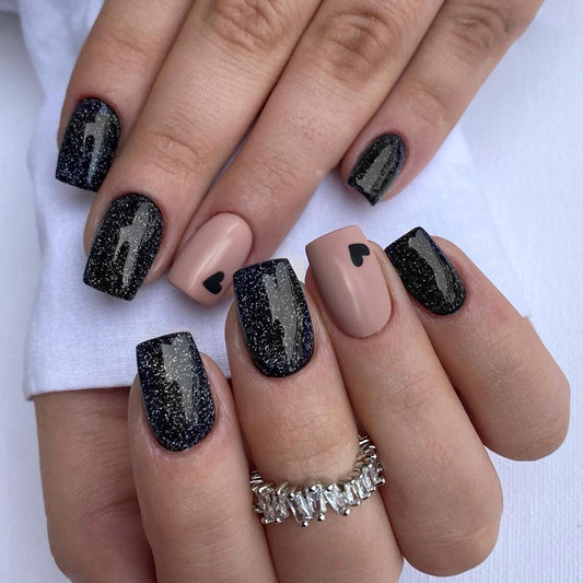 A person's hand showcasing press-on nails with a short square shape. The nails are black with glitter, and some are adorned with a silver design.