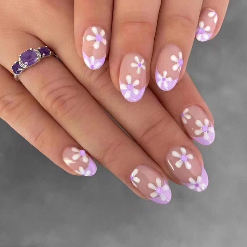 A set of multicolored floral design press-on nails, with a prominent purple and white color scheme, displayed on a hand.