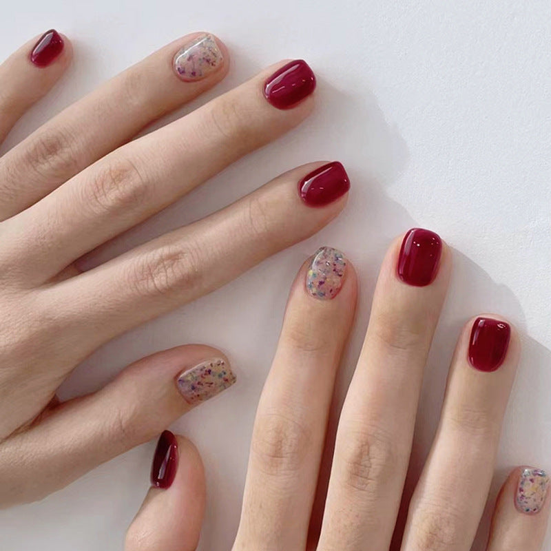 A hand with press-on nails featuring a red color and a square, short nail shape, with some nails having a glittery design.