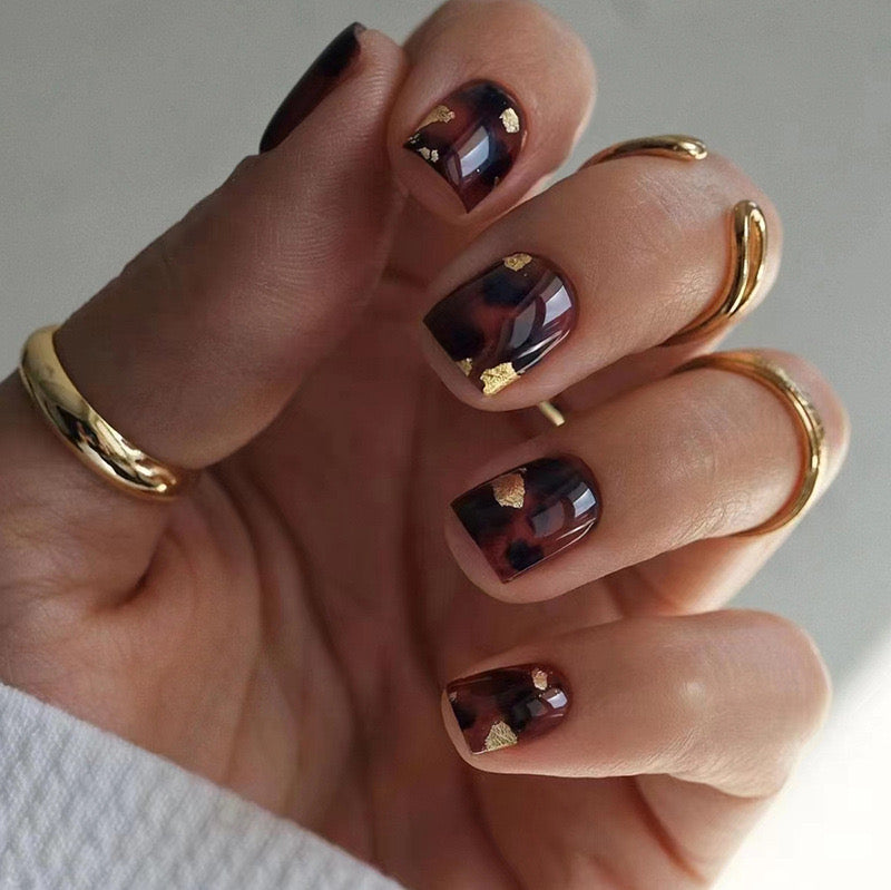 A hand with maroon press-on nails featuring gold flakes and a gold ring on the finger.