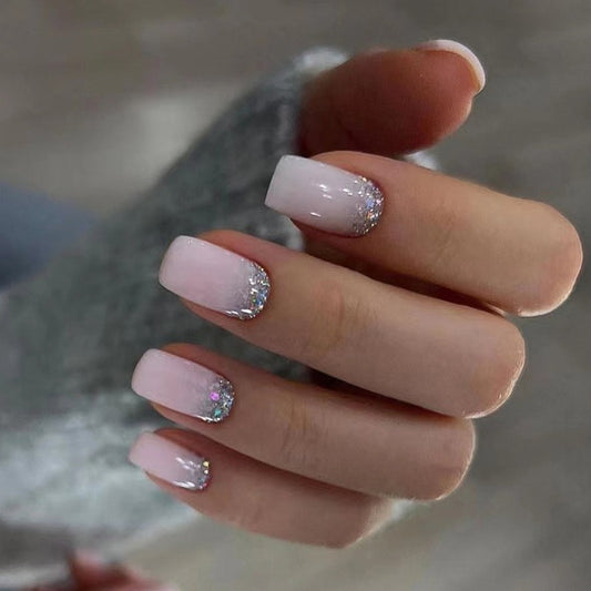 A hand showcasing press-on nails with a short square shape. The nails have a pink base with glitter accents on the tips.