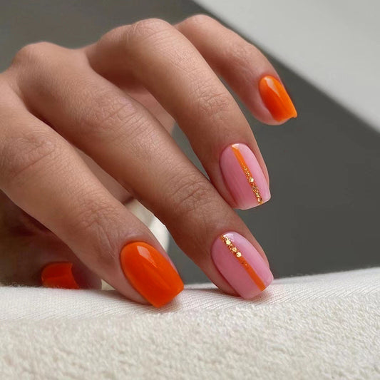 A hand with orange press-on nails with a pink tip and gold glitter accent