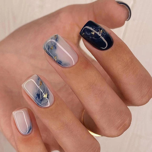 A hand showcasing press-on nails with a blue marble design and metallic gold accents.