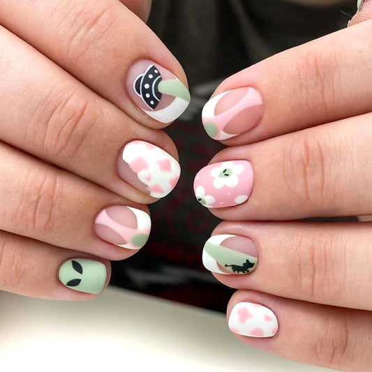 A person's hands showcasing press-on nails with a green, pink, and white color scheme, featuring cute patterns including what appears to be a flying saucer and eyes.