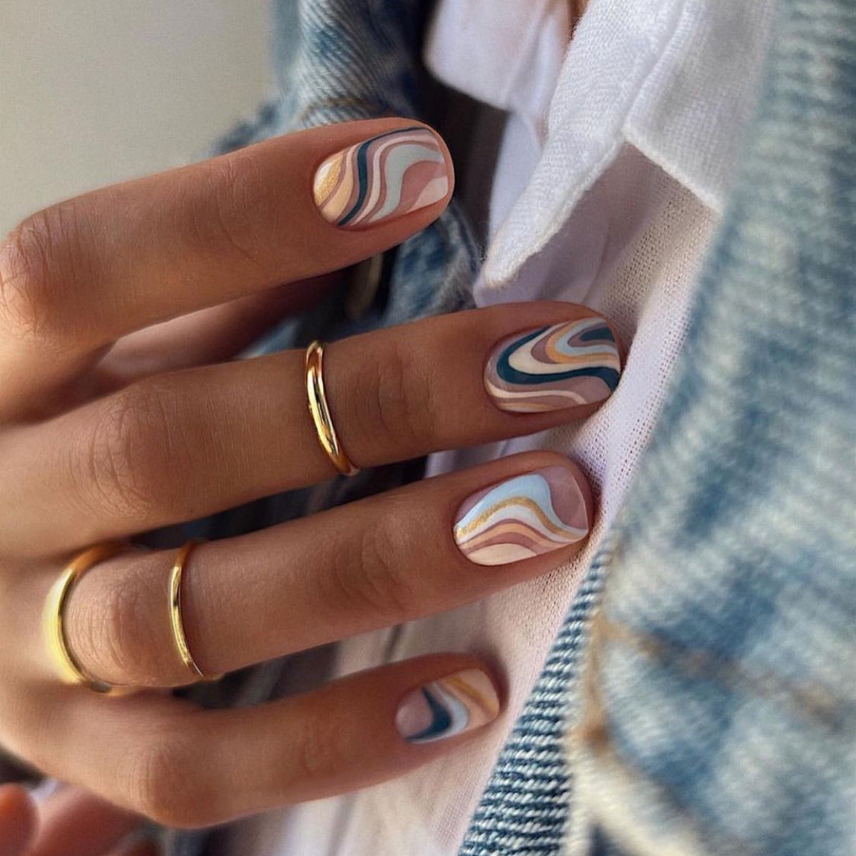 Hand with marbled nail art wearing gold rings, against a plaid fabric background