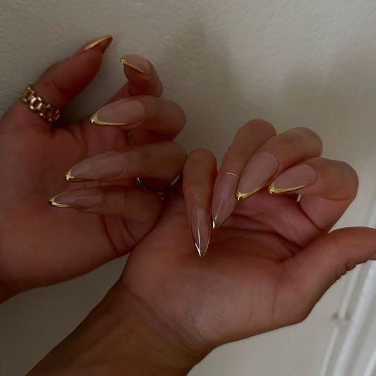 Close-up of hands with long, pointed nails and gold nail tips against a neutral background.