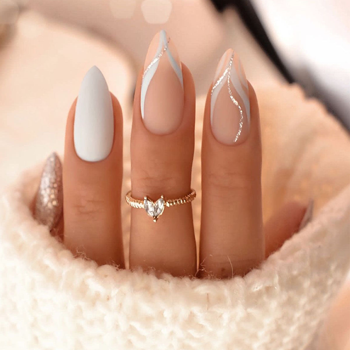 A set of long almond neutral and white press-on nails with silver glitter accents, displayed on a hand partially covered by a white sweater.