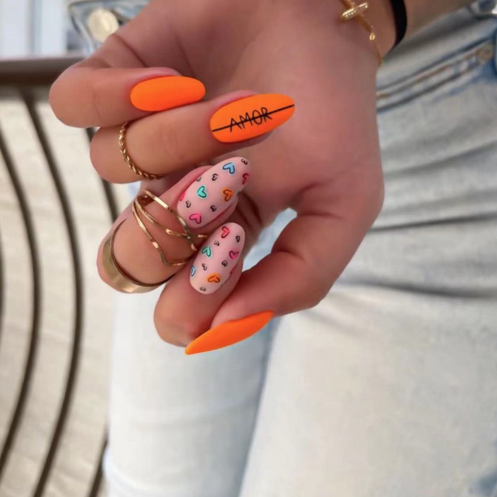 A hand with vibrant orange press-on nails with heart accents, held by a person with gold-colored rings on their fingers.