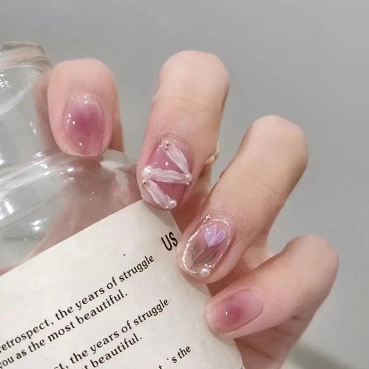 A hand with short squoval pink jelly press on nails with a floral design, holding a glass bottle.