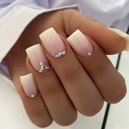 A hand showcasing press-on nails with a gradient design transitioning from pink to a solid white tip, adorned with small multicolored rhinestones.